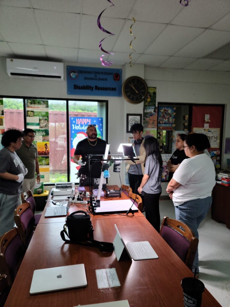 A bald man in a black t-shirt gestures over several pieces of technology including a flatbed scanner and copy stand, surrounded by a group of people listening to him.