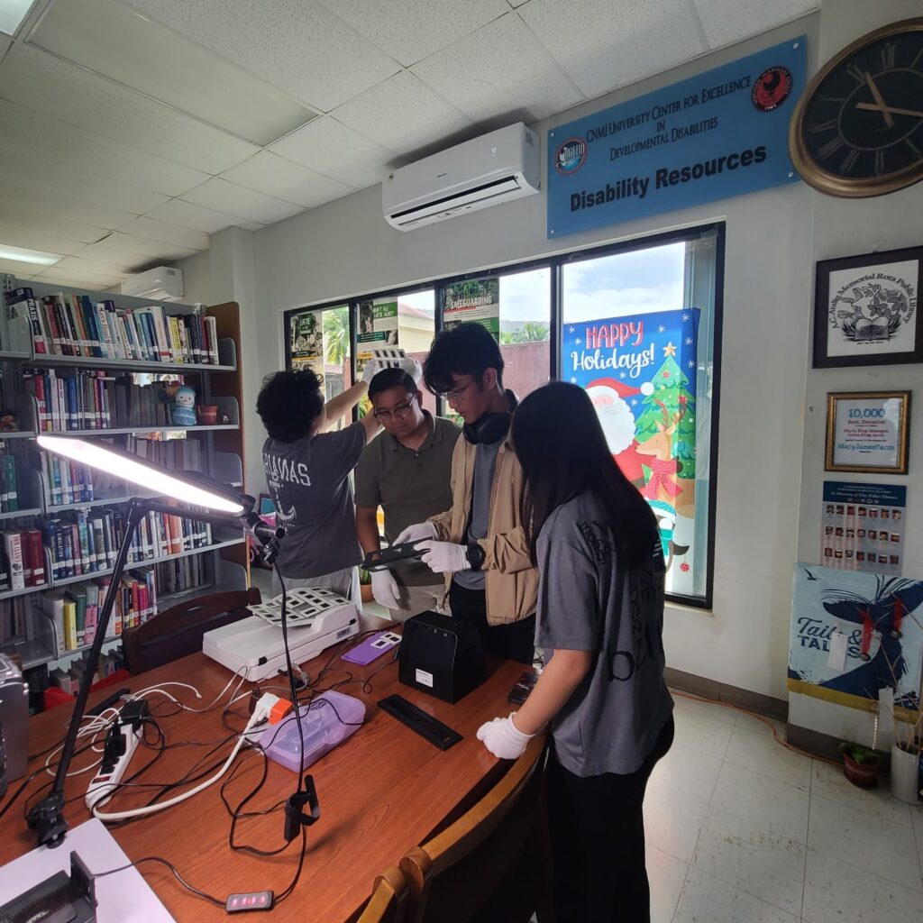 Four people stand over a flatbed scanner and other digitization technology at the end of a table in a library while examining photo slides.